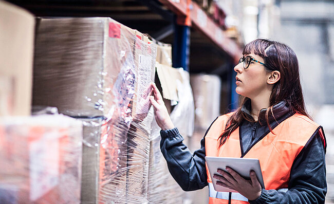 Une femme portant un gilet de haute visibilité devant un rayonnage d’entrepôt contenant des cartons